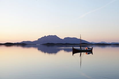 Norlandsbåt ligger på blikkstille sjø. Fjell og holmer speiler seg i sjøen i bakgrunnen