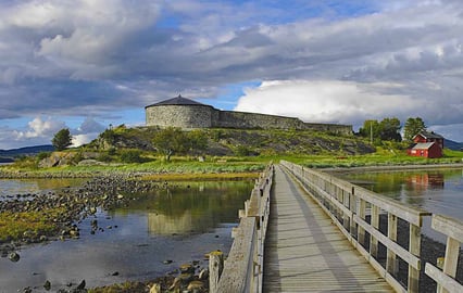 En rund steinbygning med tretak og en mur som henger fast til bygningen ligger på en øy. Det er en trebro som går over til øya