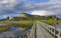En rund steinbygning med tretak og en mur som henger fast til bygningen ligger på en øy. Det er en trebro som går over til øya