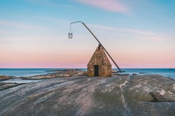 En kopi av et gammelt vippefyr i stein står på et svaberg. Rosa solnedgang i bakgrunnen
