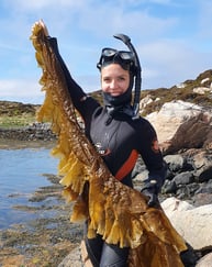 Ung, smilende dame i våtdrakt og dykkemaske holder opp en stor brunalge