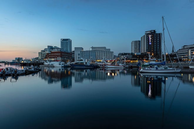 Kveldsbilde av en rekke høye hus som ligger langs kaia. I forgrunnen er det fiskebåter, små ferger og seilbåter