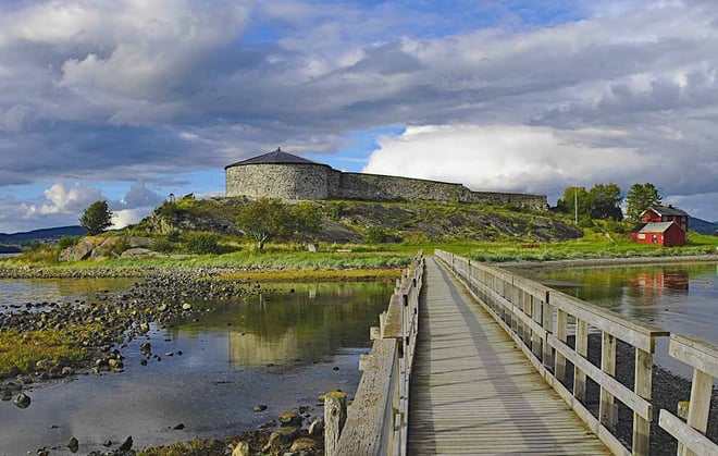 En rund steinbygning med tretak og en mur som henger fast til bygningen ligger på en øy. Det er en trebro som går over til øya