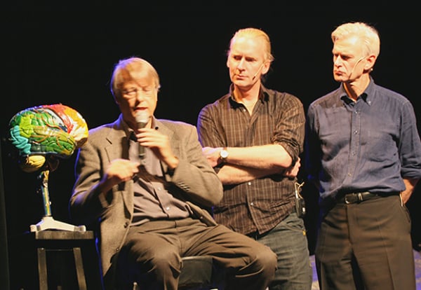 tre mannlige skuespillere på en mørk scene ved siden av en fargerik modell av en menneskelig hjerne opphøyet på et stativ.