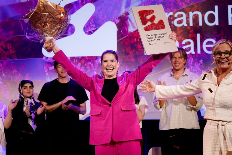 Person i rosa dress på scenen holder opp blomster og et diplom med teksten «Vinner av nasjonal finale i Forsker Grand Prix», mens personer i bakgrunnen applauderer.