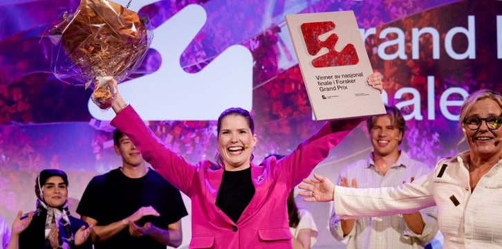 En glad kvinne i rosa blazer holder opp en blomsterbukett og diplomet «Vinner av nasjonal finale i Forsker Grand Prix», mens flere personer applauderer i bakgrunnen.