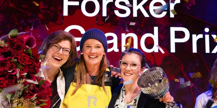Tre personer poserer med blomster og en stor glasspokal foran en skjerm med teksten «Forsker Grand Prix», mens konfetti faller rundt dem.