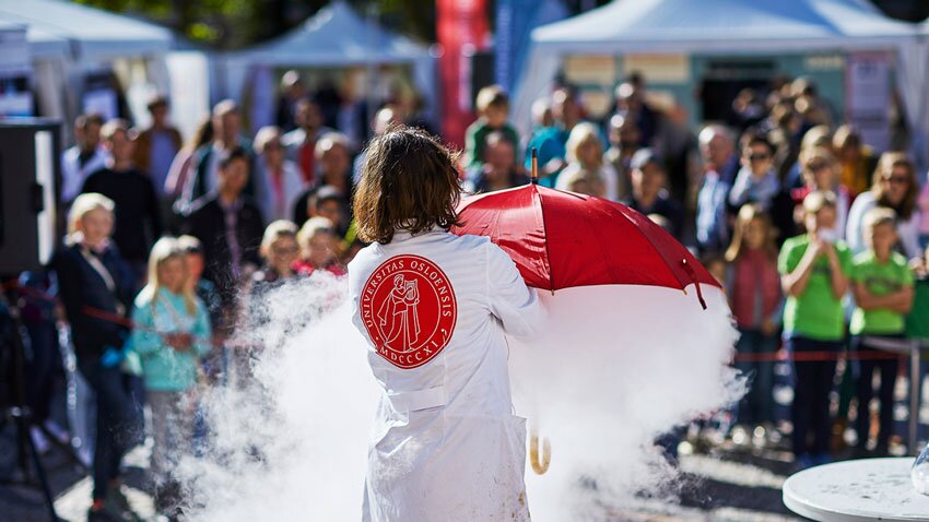 Fra Forskningstorget i Oslo - person med hvit frakk med ryggen til holder en rød paraply med hvit røyk under. Publikum uskarpt i bakgrunnen.